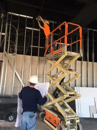 A man with blueprints and another on a scissor lift working on a building